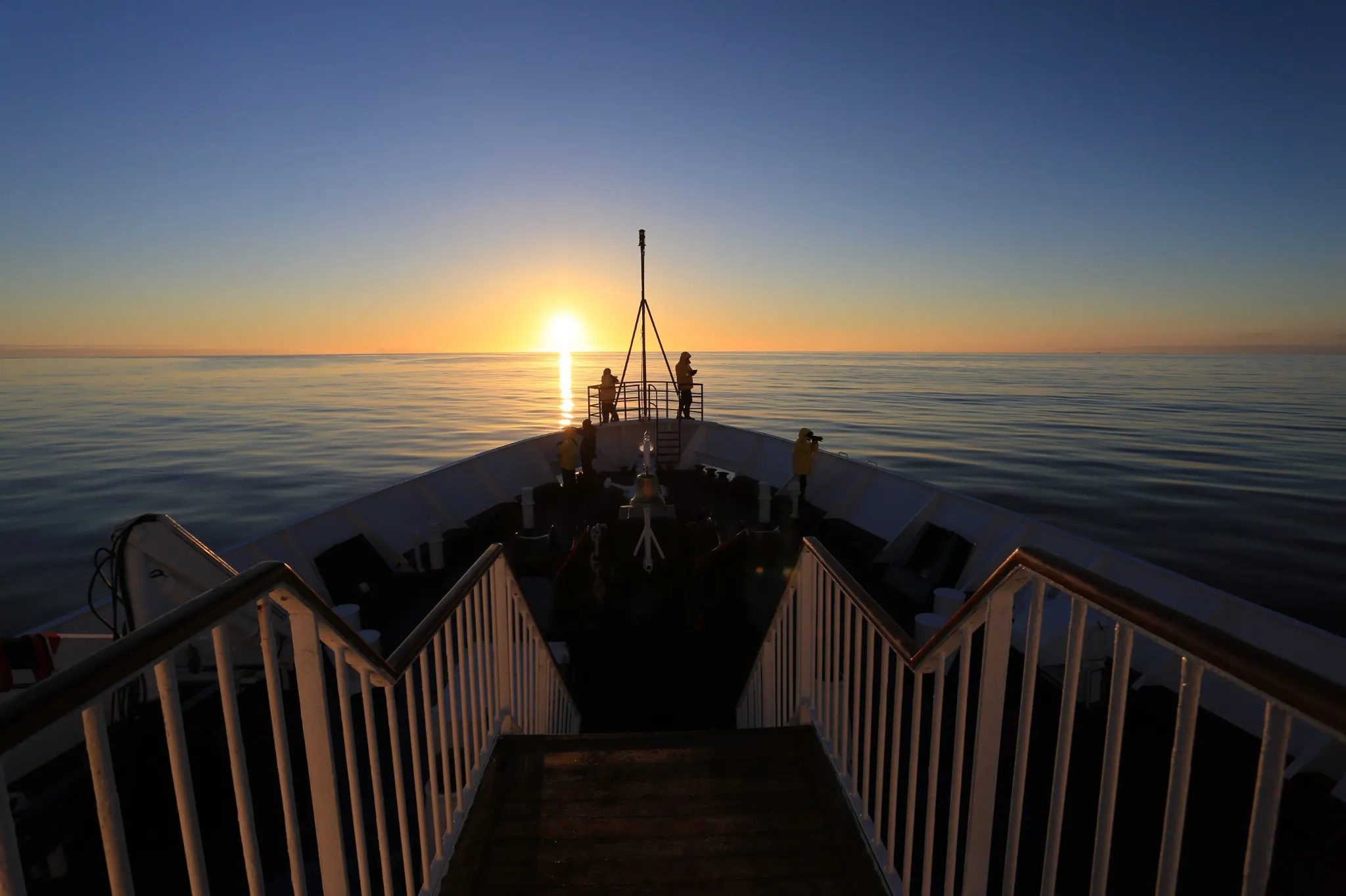 Expedition guests in yellow jackets on the bow of a ship, silhouetted against the rising sun over a calm, glassy sea