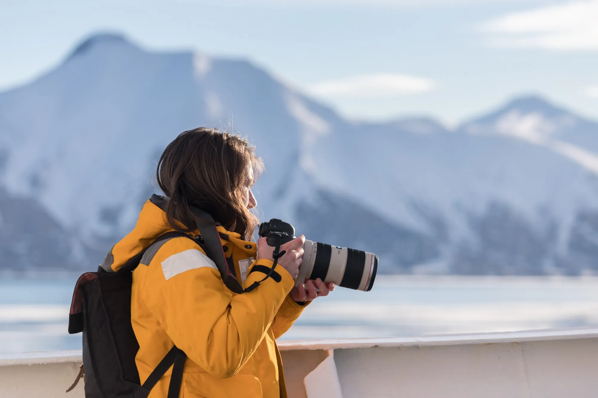 A guest in a yellow parka photographing snow-covered mountains from the ship’s deck