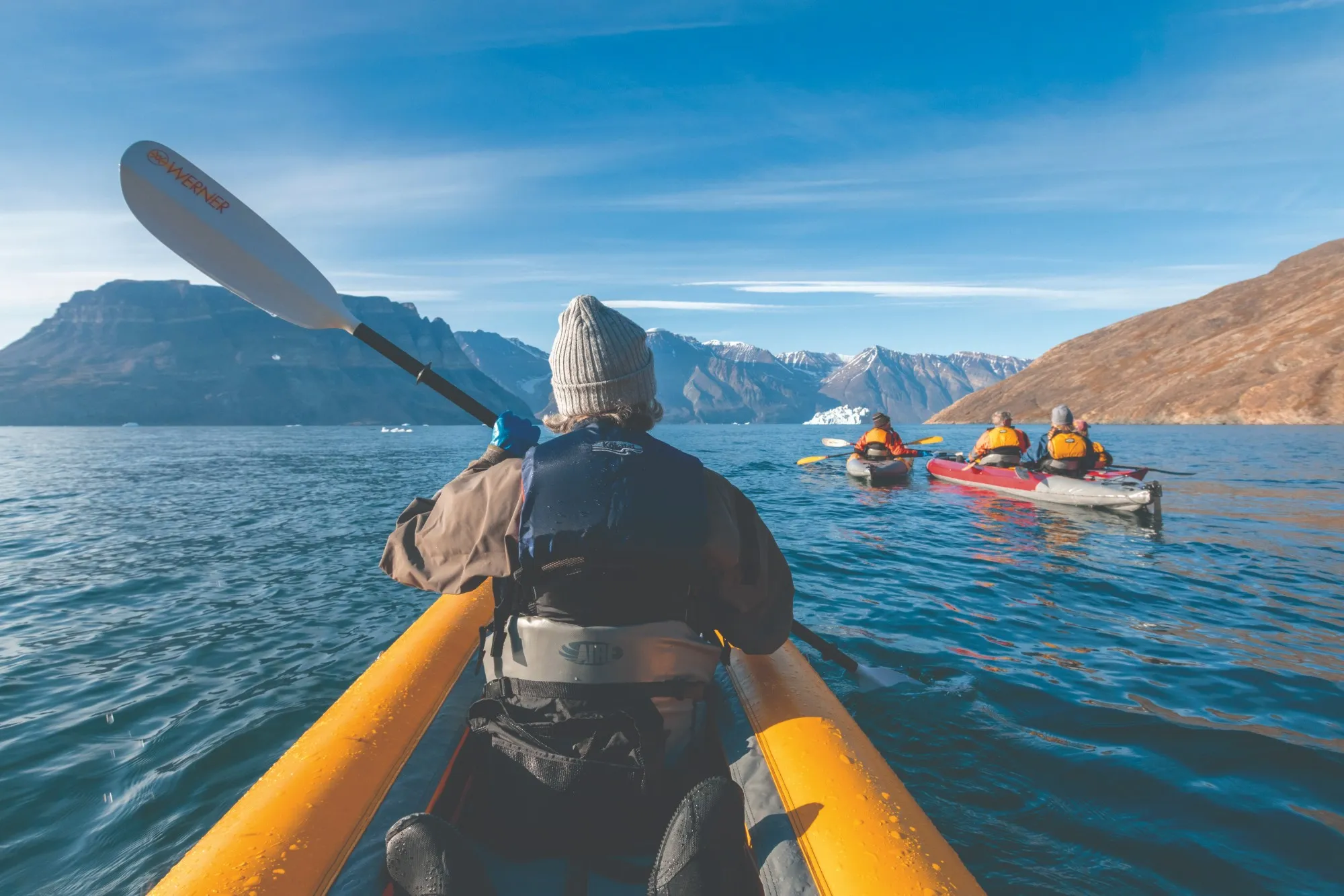 View from a sea kayak following others up a calm fjord towards mountains and an iceberg