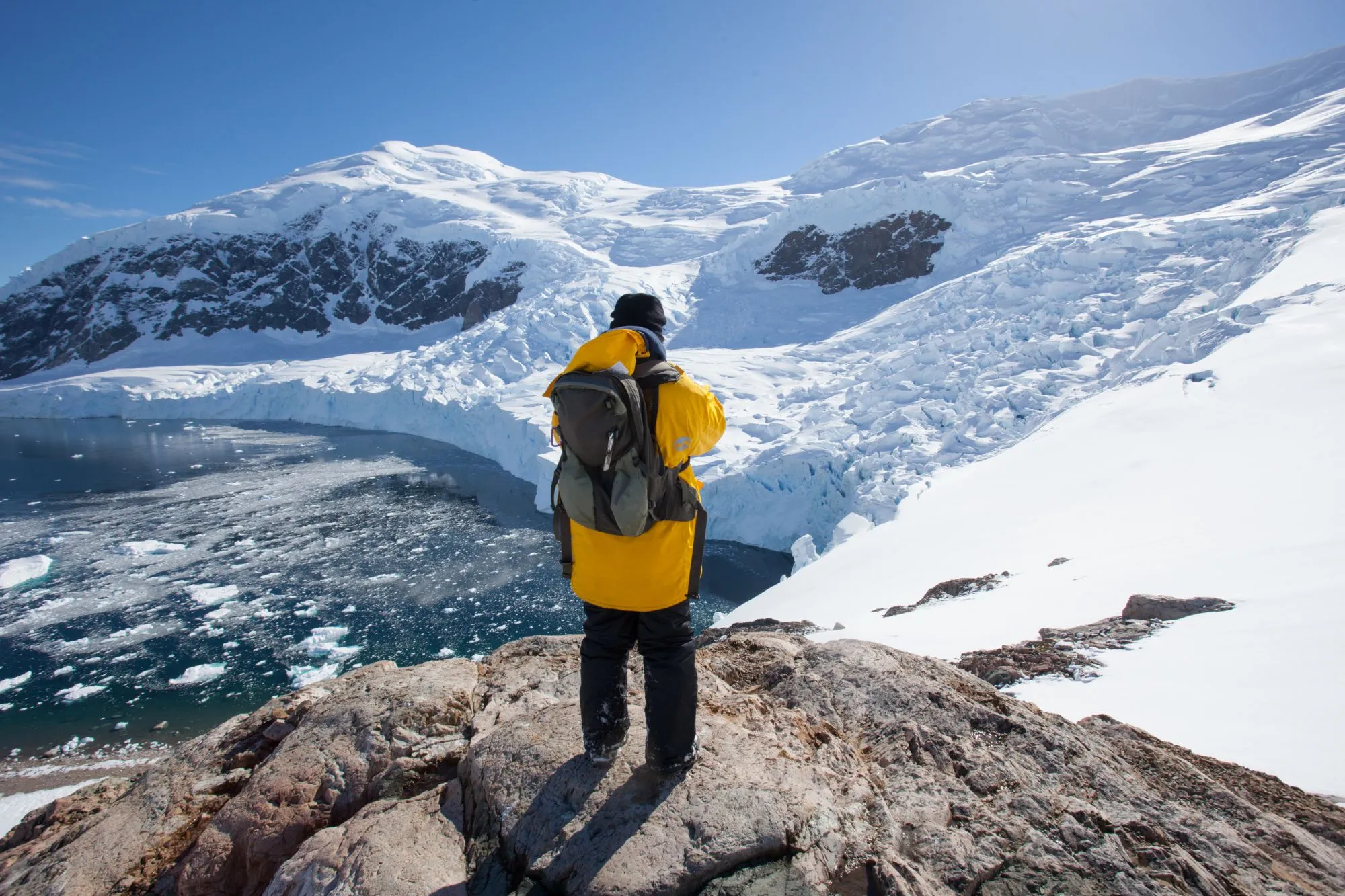 A guest in a yellow parka looks down over a glacier and an ice-filled bay