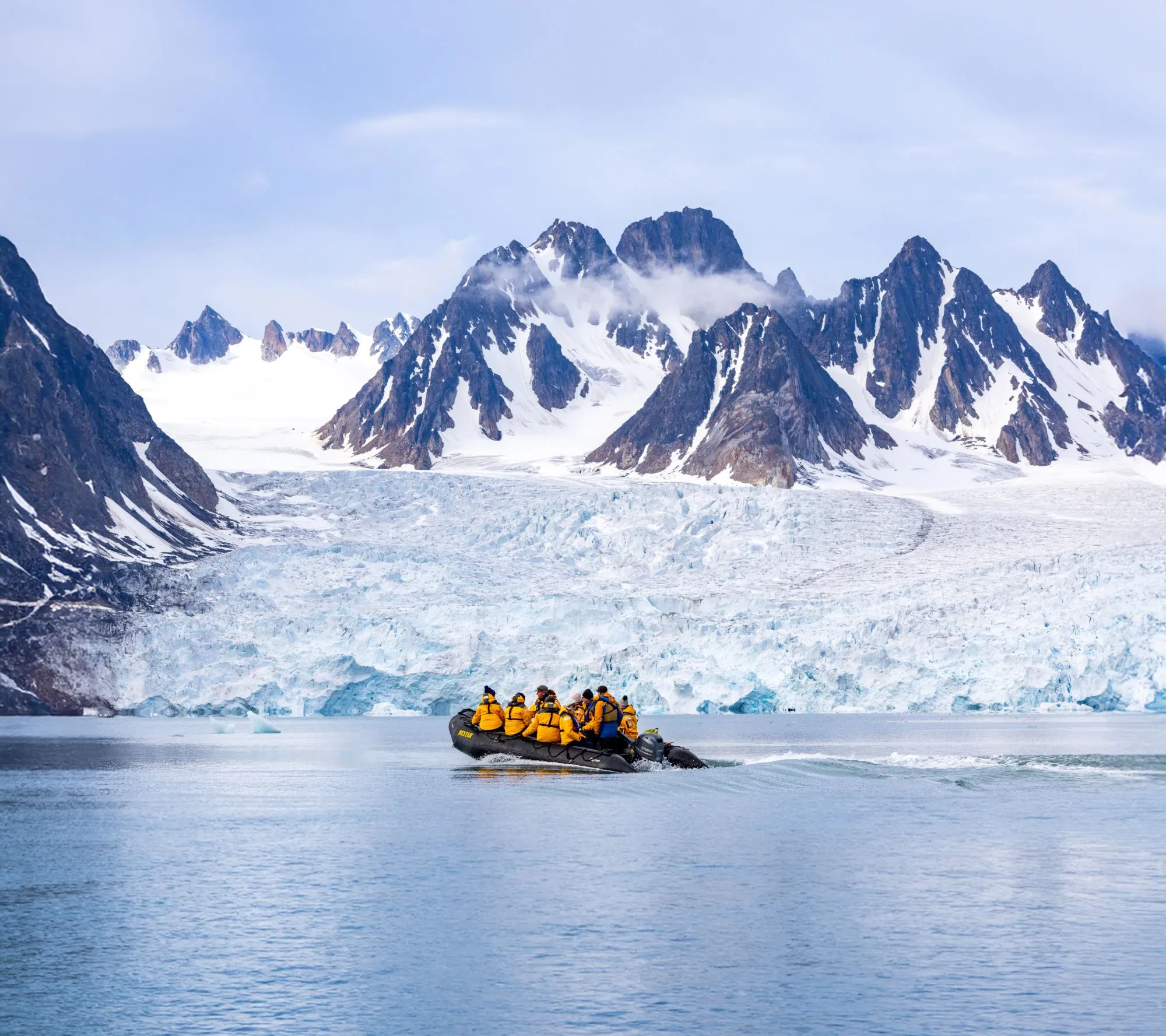 A zodiac of guests in yellow parkas crossing calm water in front of a vast glacier and jagged peaks