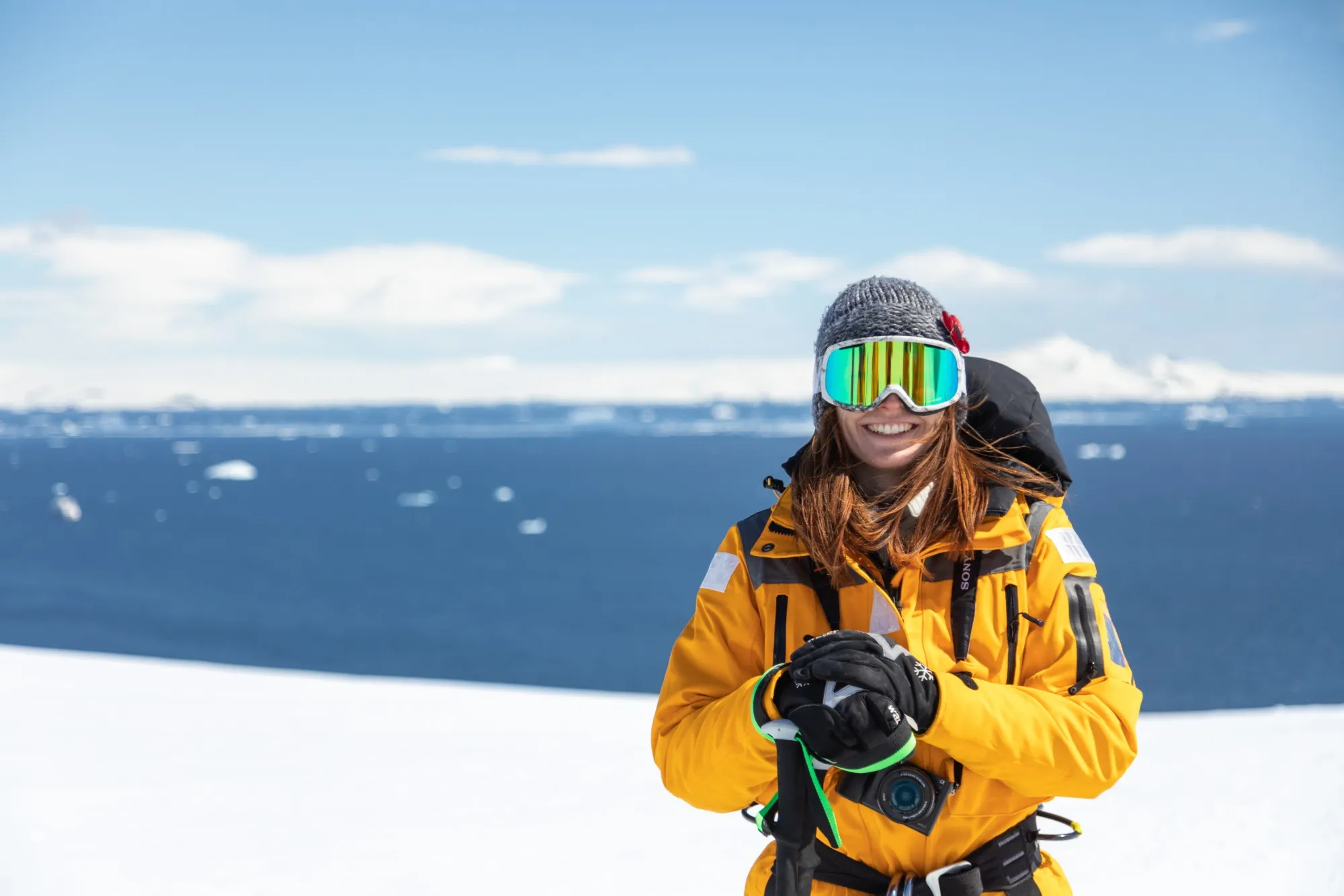 A smiling guest in goggles and a yellow parka on a snowy summit above the sea
