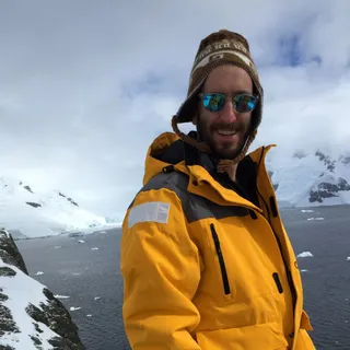 Mike Abbott smiling in a yellow expedition jacket on a snowy ridge in Antarctica, with sea ice and glaciers behind