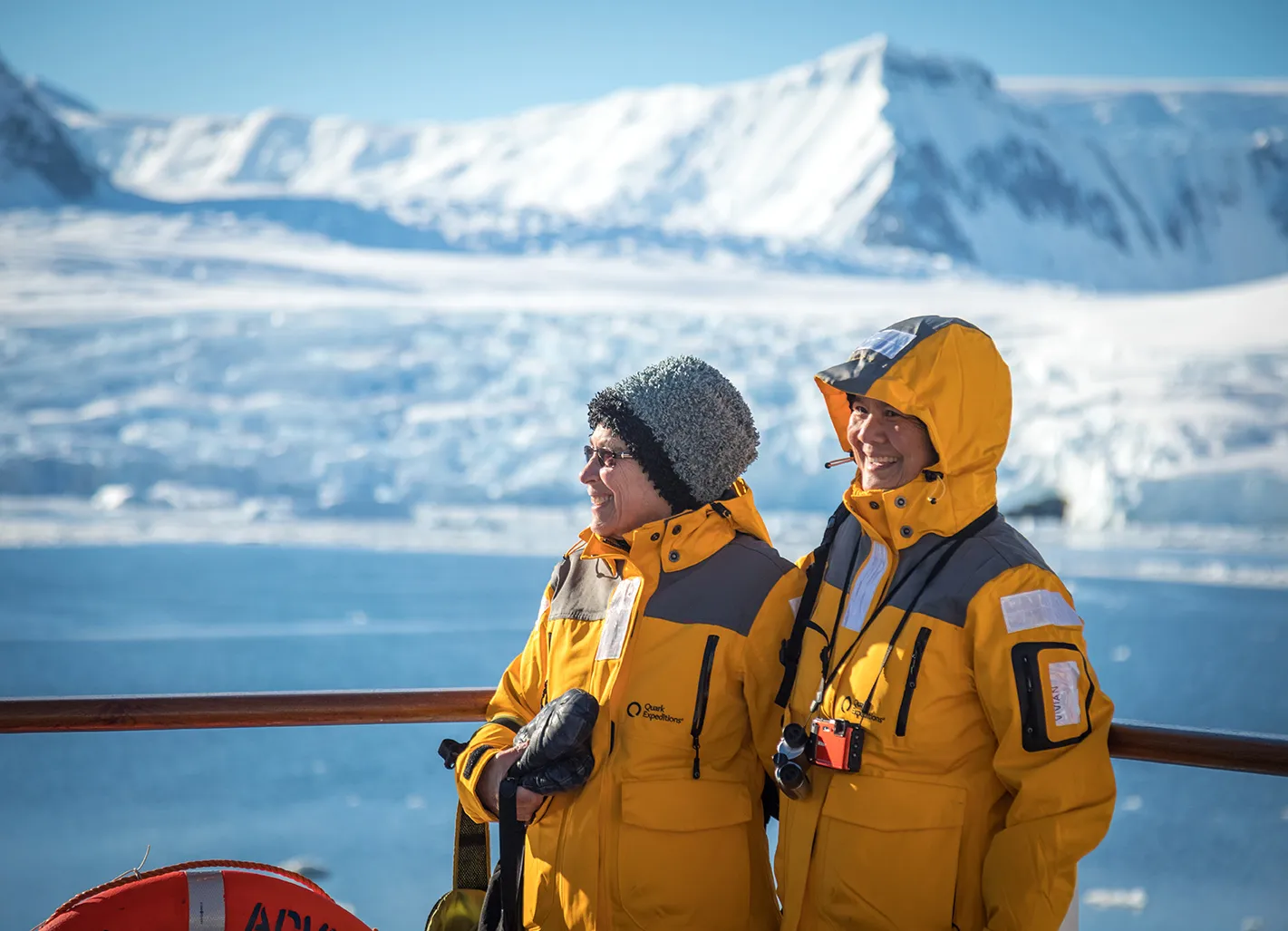 Two smiling guests in yellow parkas at the ship rail, a glacier behind them