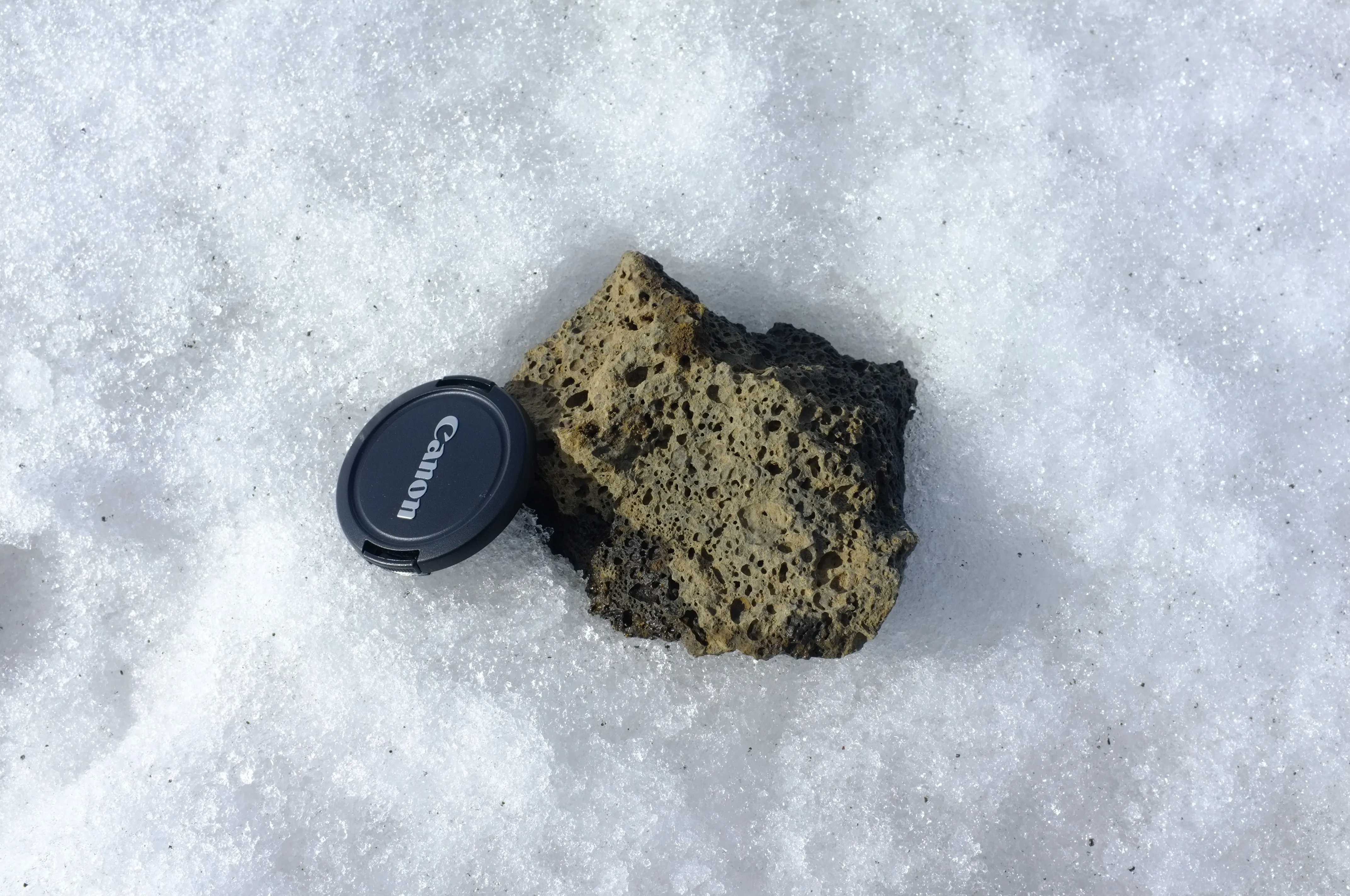 A dark, porous rock full of small holes lying on coarse snow beside a Canon lens cap for scale