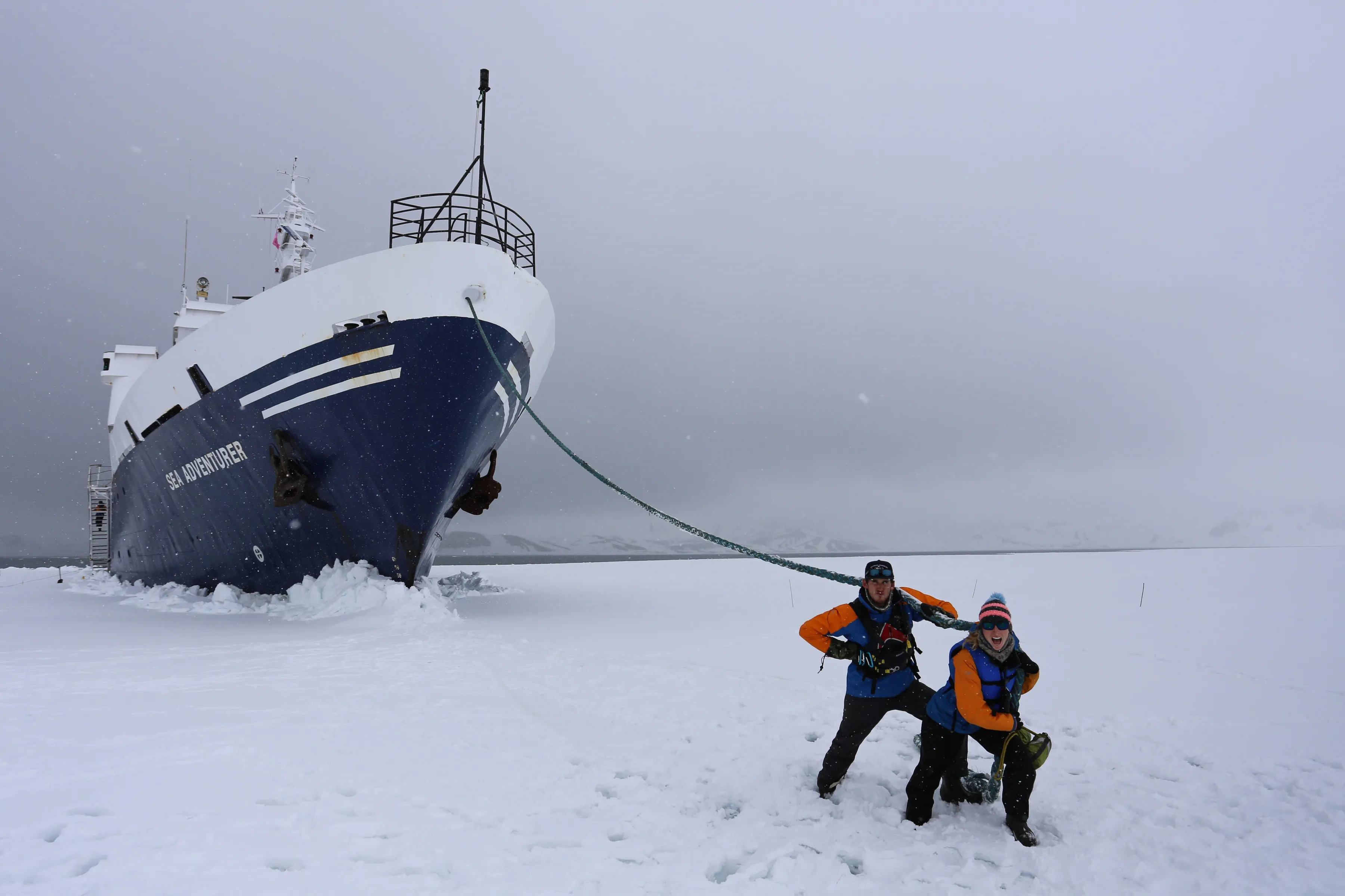 Will and Liz, in orange and blue expedition jackets, mime hauling on a thick mooring line from the bow of the blue-hulled Sea Adventurer, held in snow-covered sea ice