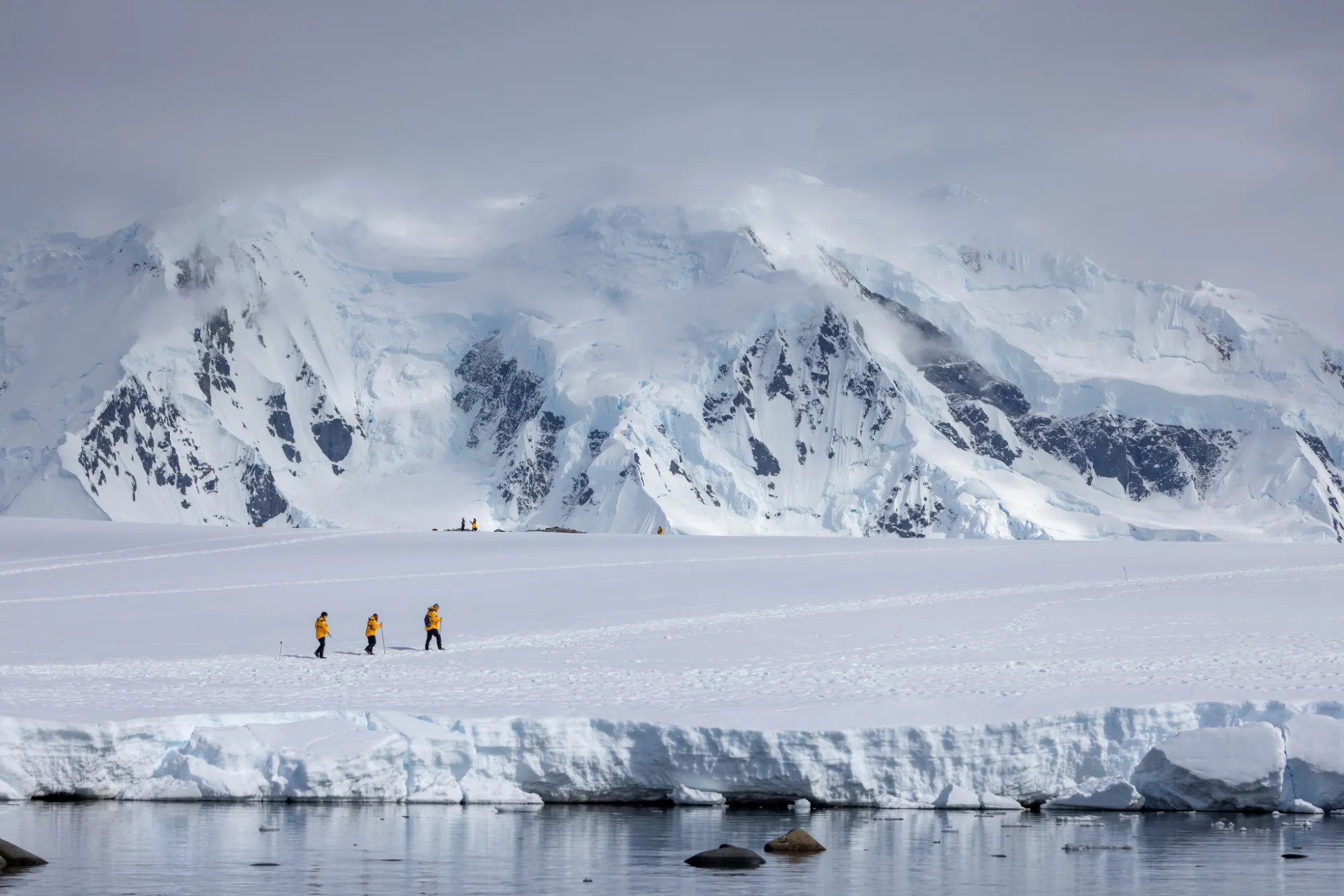 Three guests in yellow parkas hiking across a snowfield beneath cloud-wrapped glaciated peaks