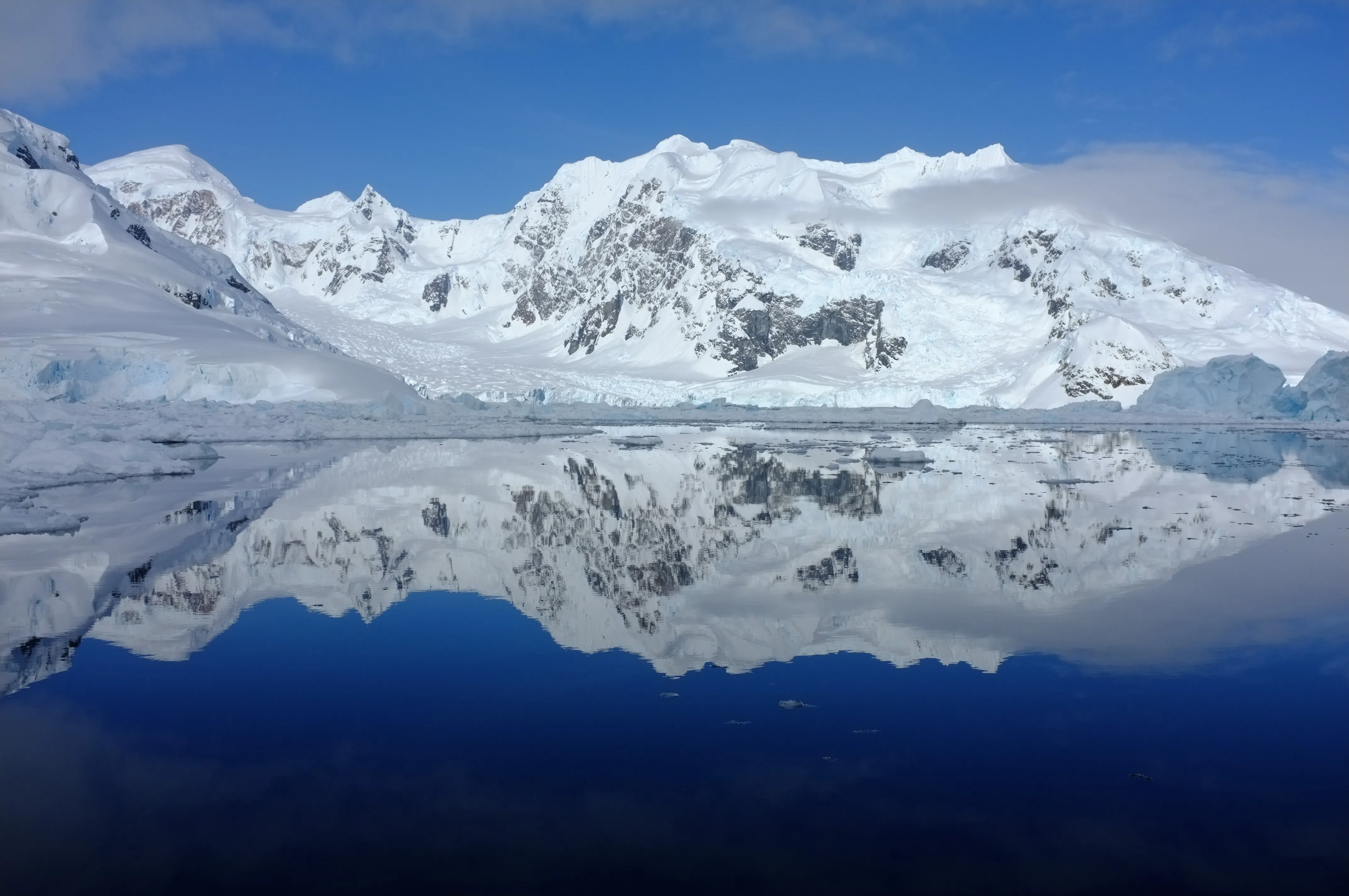 Snow-covered mountains and glaciers mirrored in perfectly still, deep-blue water under a clear sky