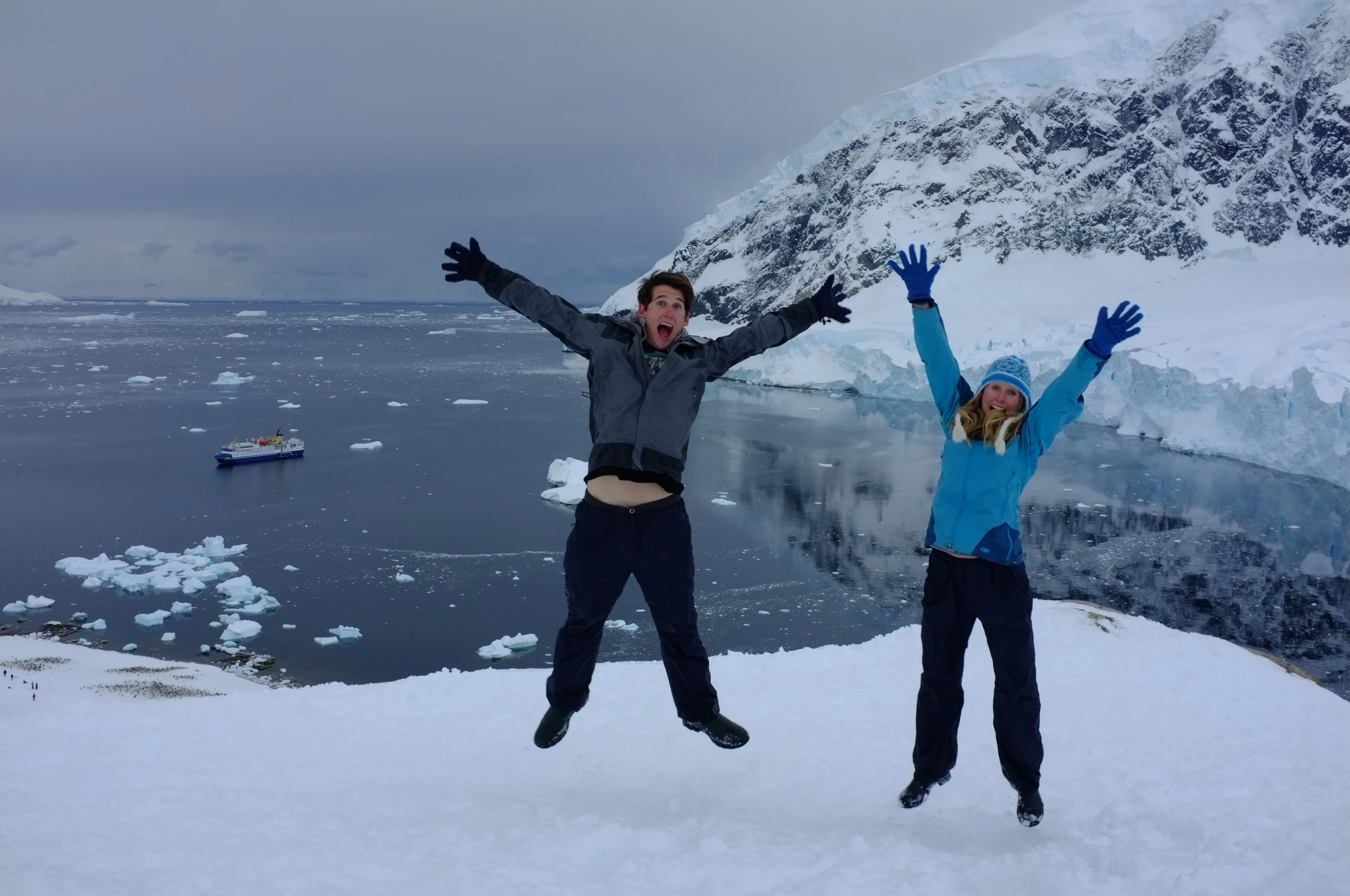 Will Abbott and Liz Teague jump with their arms outstretched on a snowy hilltop, with a ship anchored among floating ice in the bay far below
