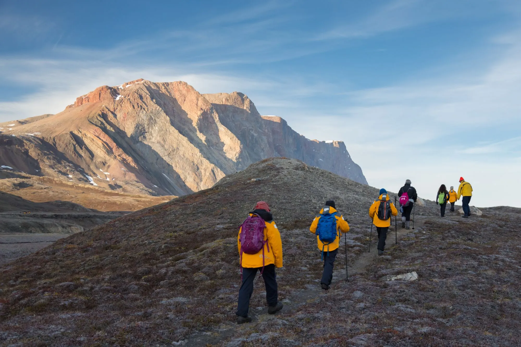 A line of guests in yellow parkas hiking a tundra ridge towards a sunlit red mountain