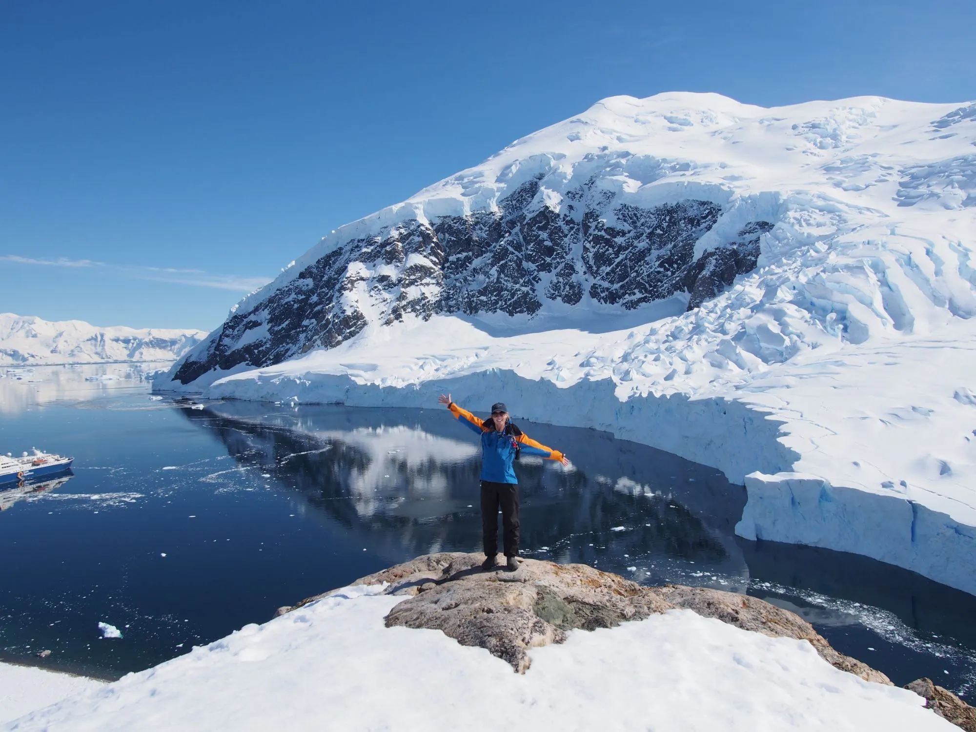 A person in a blue and orange jacket stands with arms outstretched on a rocky, snowy viewpoint above a calm bay, with a glacier front, a snow-covered mountain and an expedition ship below