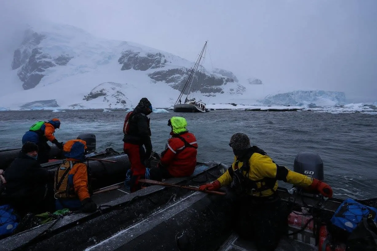Expedition crew in Zodiacs on choppy grey water in falling snow, with a sailing yacht heeled over on the rocks of a snowy island behind them
