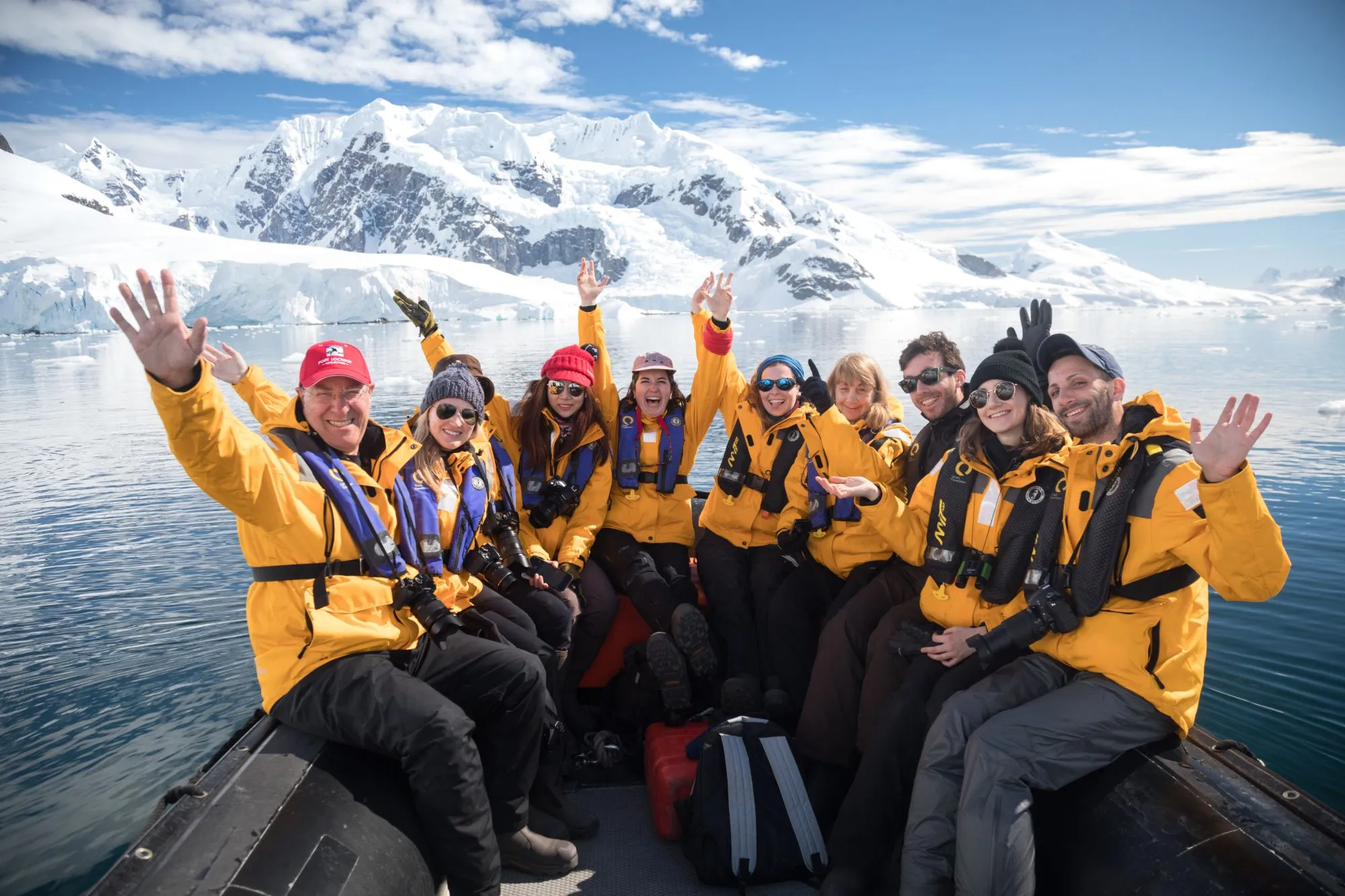 A zodiac full of guests in yellow parkas waving, glaciated mountains behind
